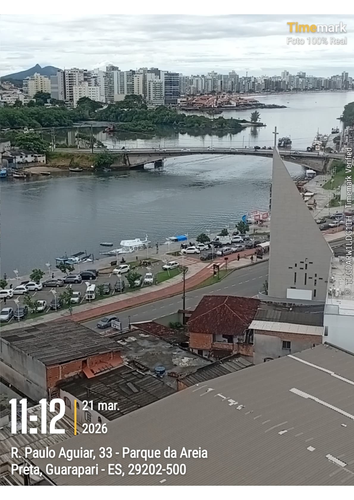 Vista panorâmica elevada mostrando rio, ponte, ruas movimentadas, construções urbanas e prédios ao fundo.